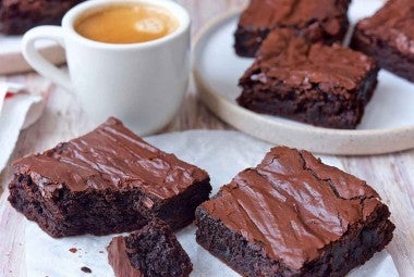 Several fudge brownies with shiny, crackly tops, sliced next to a cup of coffee