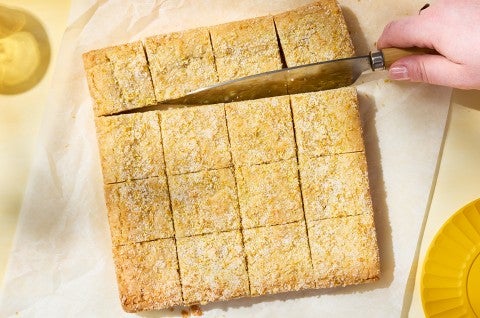 Slicing Alex Roberts Lemon Citrus Blondies on parchment paper atop a counter. - select to zoom