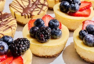 The side of golden yellow Mini Cheesecakes topped with fresh berries and others with chocolate drizzle.
